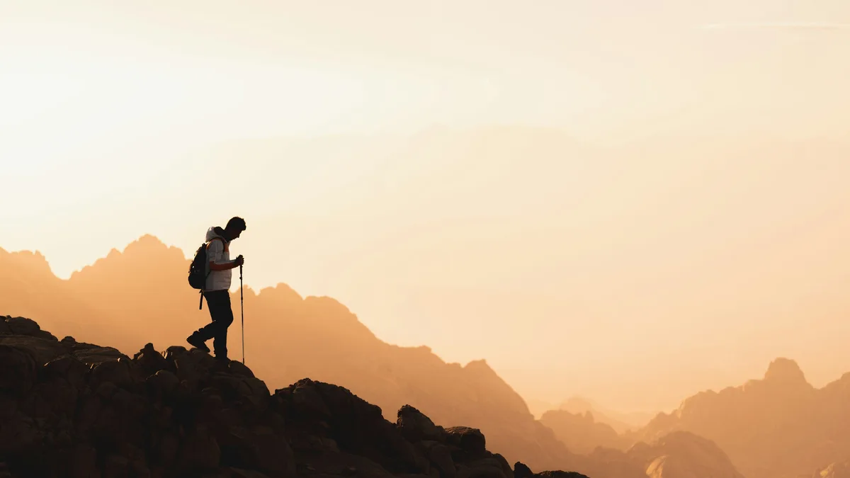 A man alone on a mountain ridge at sunset, hiking pole in hand, silhouetted against golden light