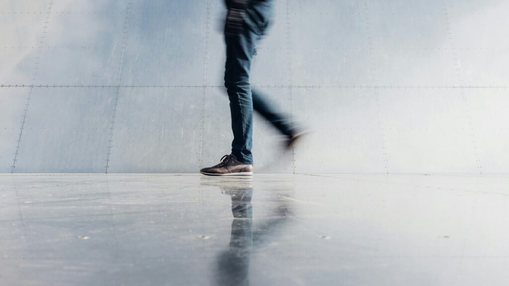 Person walking past a pale wall in dark trousers and shoes