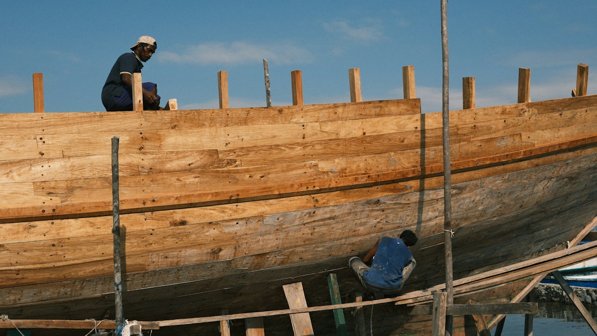 Two people beside a large wooden boat hull under construction on land