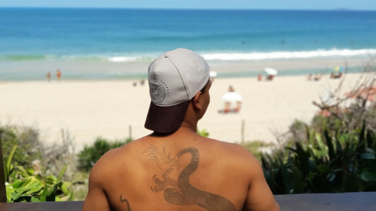 Man with a curved dragon tattoo extending diagonally across his upper back while looking toward a beach