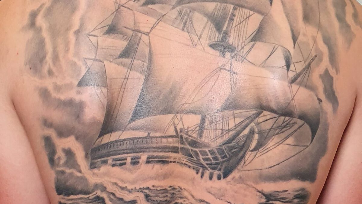 Black and grey sailing ship tattoo across a man's back, with dark storm clouds and choppy waves