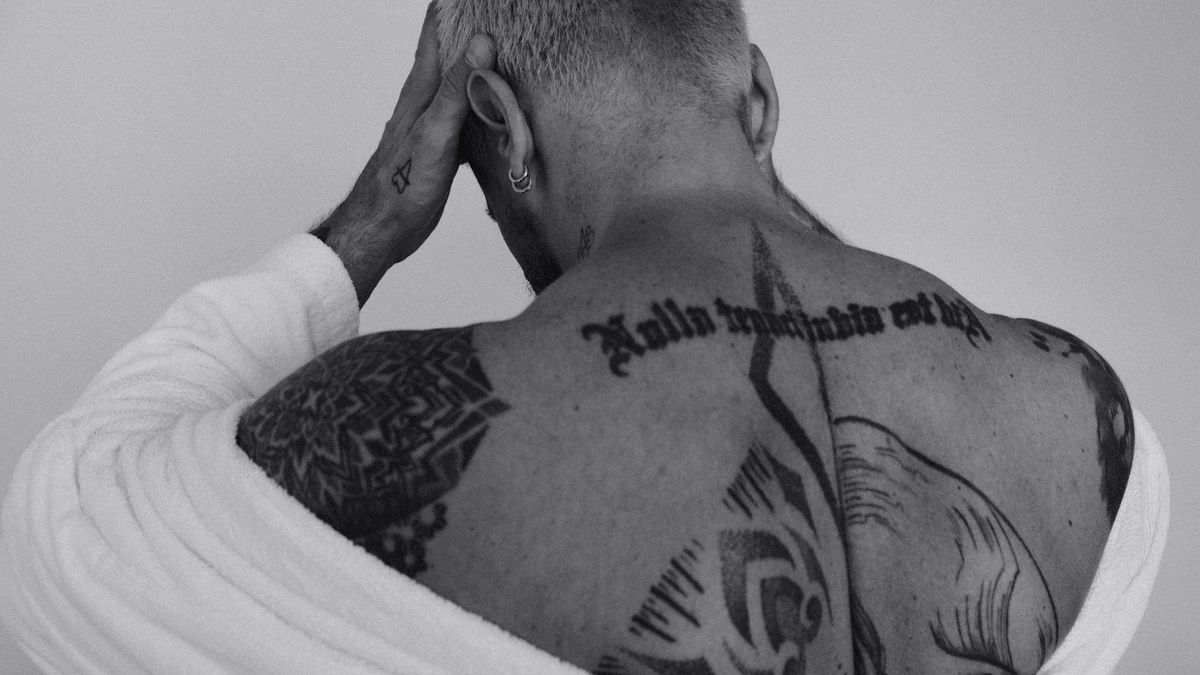 Black and white photograph of a man with several differently styled upper-back tattoos, including lettering and geometric artwork