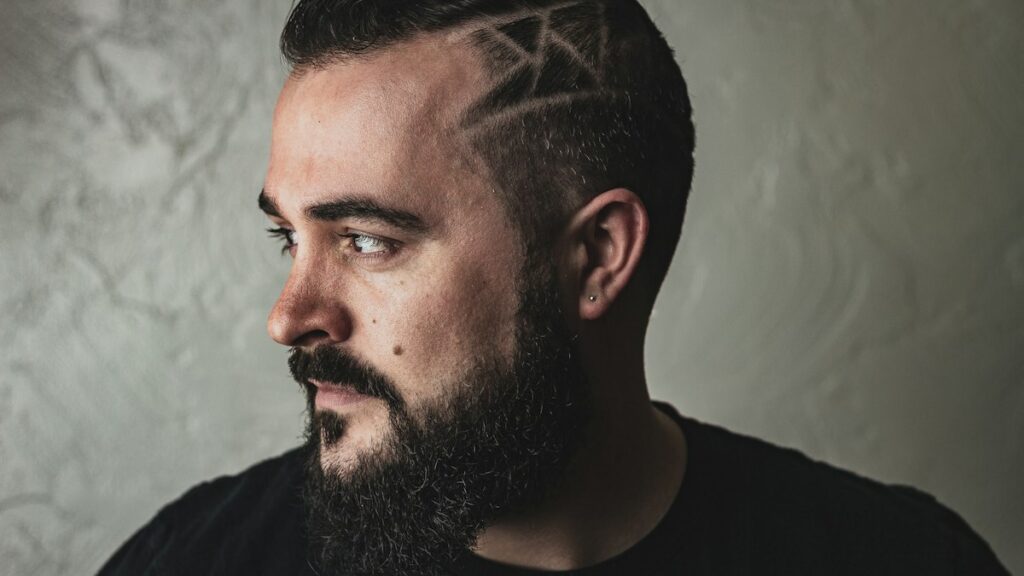 Man with a full dark beard and a patterned side haircut looking to the left