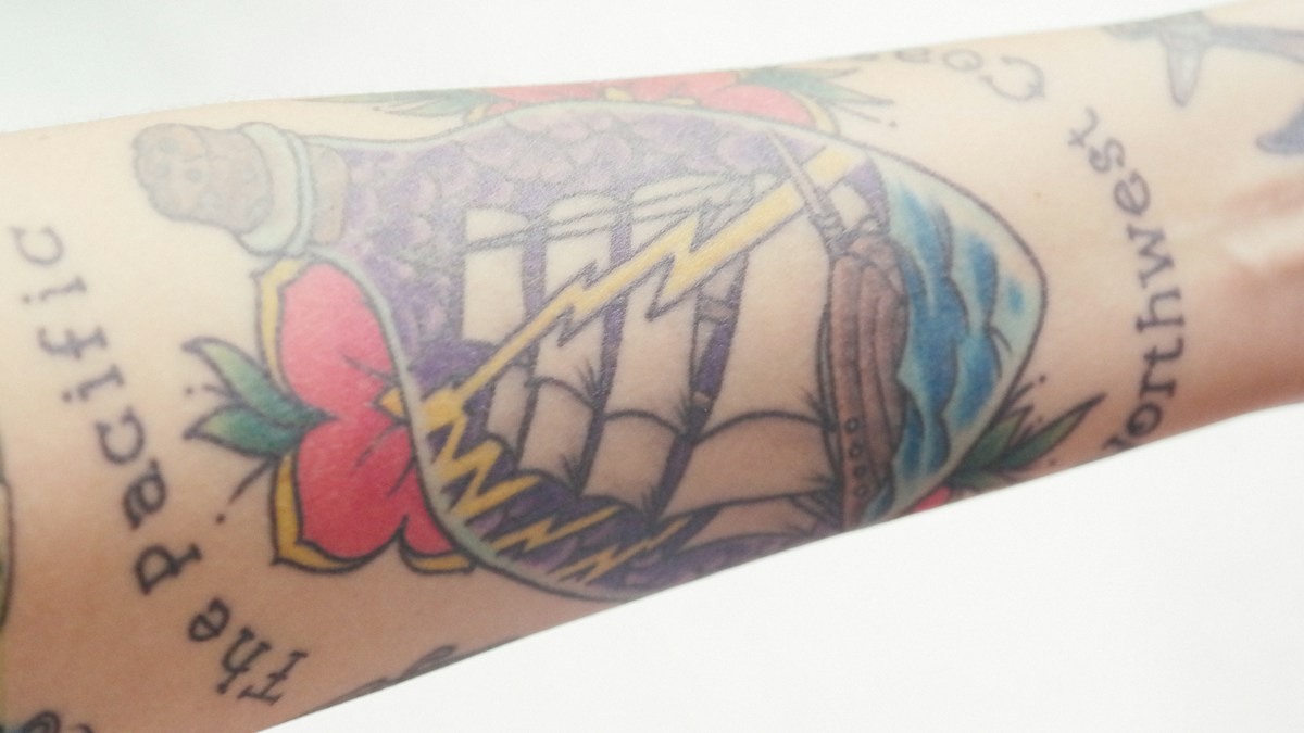 Vintage-style ship in a bottle tattoo with colorful lightning and floral motifs on a forearm