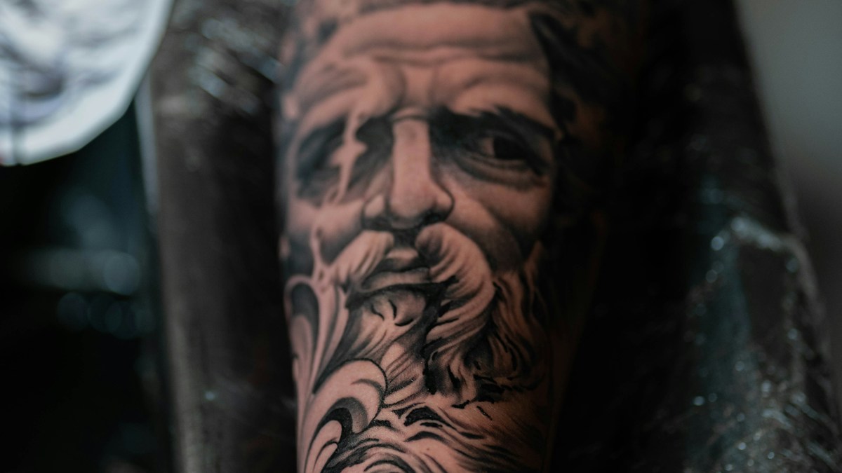 Black and grey tattoo of a bearded classical sculpture face on the forearm, showing detailed stone-like shading