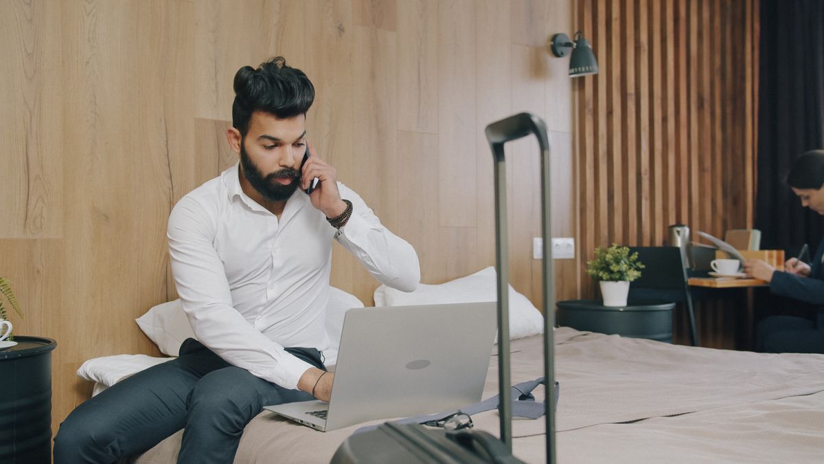 Bearded business traveler using a laptop in a hotel room with luggage nearby.