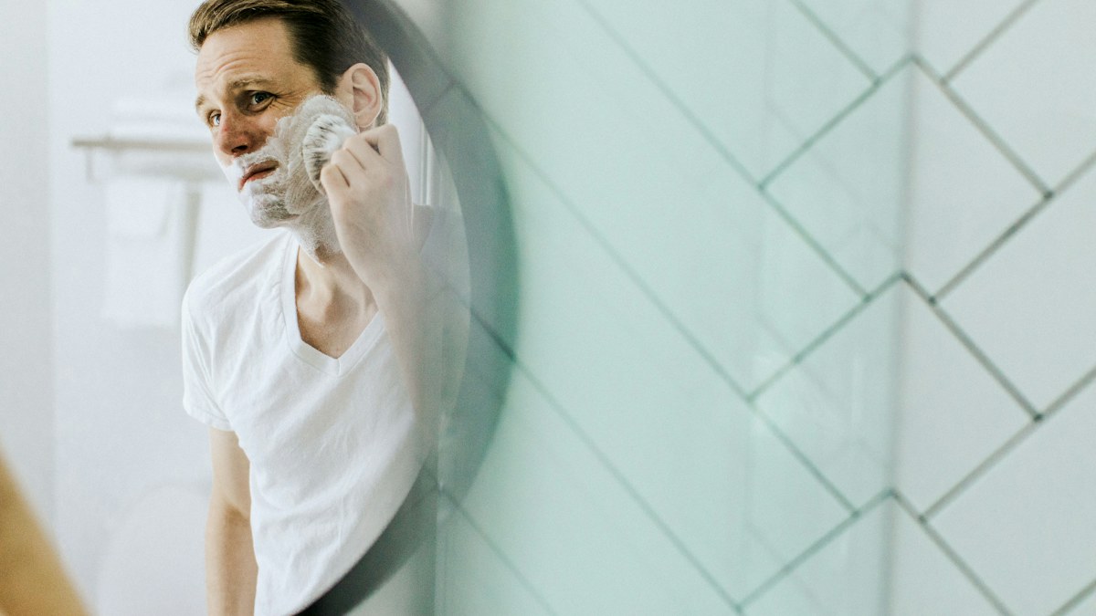 Man applying shaving lather with a brush in front of a bathroom mirror