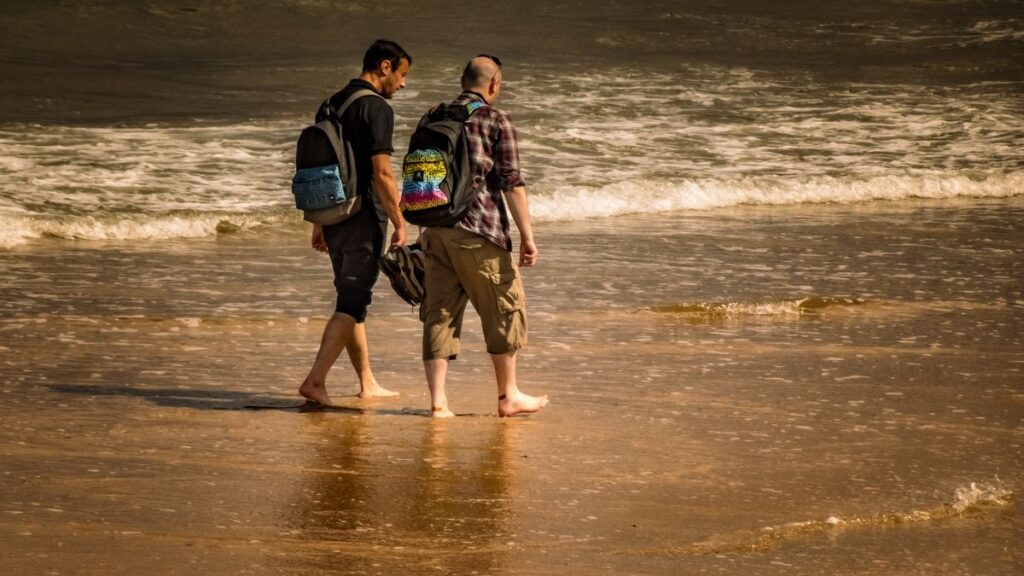 Two men carrying backpacks beside a beach.